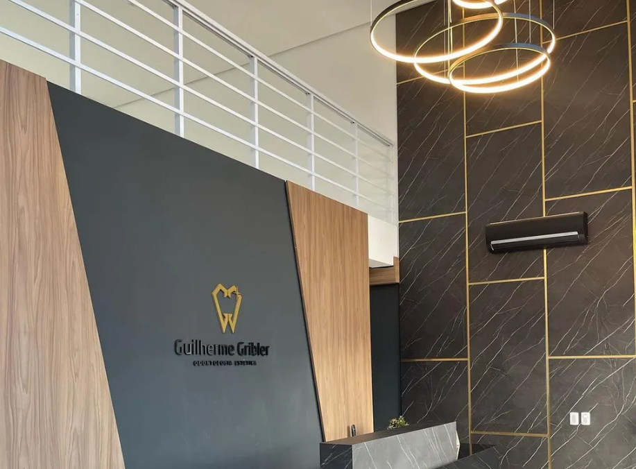 Recepção do consultório com o logotipo Guilherme Gribler em dourado, painéis de madeira, parede de mármore escuro e luminária circular